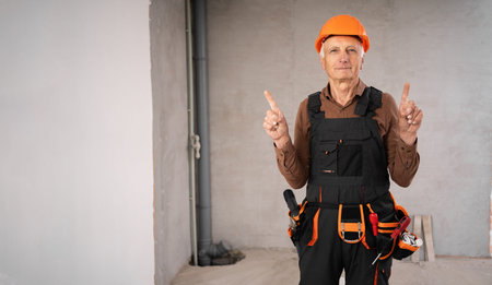 Senior builder on construction site standing wearing safety helmet pointing with thumbs-up gesture...