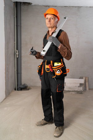 Elderly builder with tool belt holding wall measuring level and pencil looking at camera kindly. House construction and renovationの写真素材
