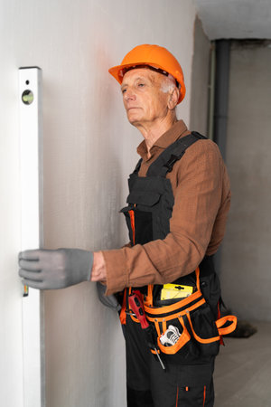 Measurement and house repair. Elderly builder or construction worker in protective hard hat checking level of wall.の写真素材