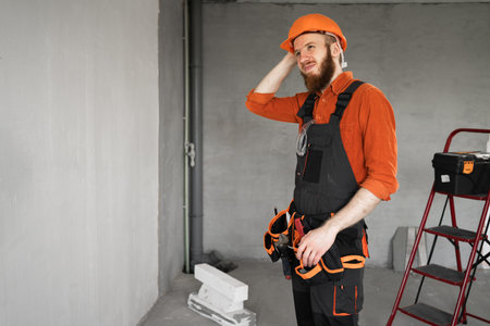Bearded repairman or builder in protective helmet and overalls having doubts with confuse face...
