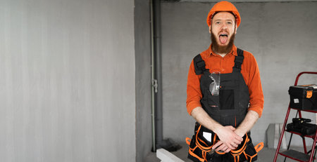 Bearded repairman or builder in protective helmet and overalls showing his tongue in the room with...