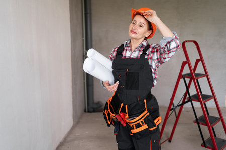 Middle aged woman builder tired after work takes off helmet holding paper project for renovation of rooms in houseの写真素材