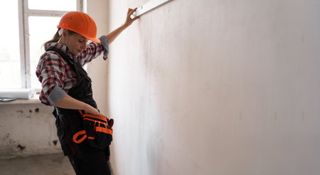 Woman civil engineer wearing hard hat and tool belt measuring wall with spirit level reaching for pencil showing focus craftsmanship precision building. home renovation processの写真素材