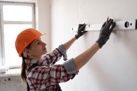 Close-up of woman builder using spirit level to measure wall and making pencil marks. Concept of...