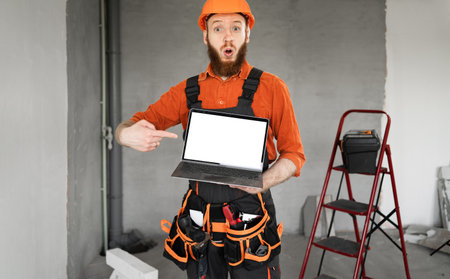 Portrait of repairman or builder in helmet and overalls holding laptop with surprised expression pointing finger at white mock up advertising spaceの写真素材