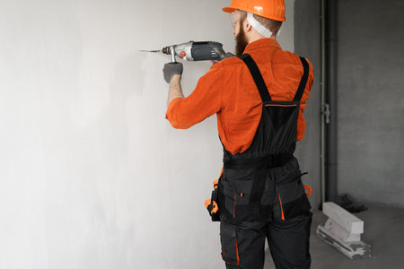 Bearded male builder in orange helmet and blue overalls working with drill perforator at...