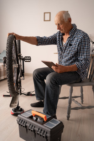 An elderly Caucasian man in a cozy apartment repairs a bicycle, studying video tutorials on a tablet. Tools are nearby, he is focused. Hobbies and active life in retirement are full of inspiration and skillの写真素材