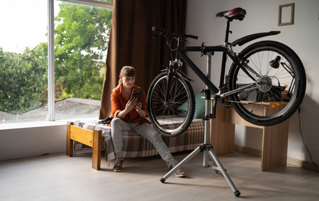 Middle aged woman repairing bicycle sitting on bed using digital tablet to search for spare parts...