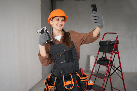 Female builder in safety helmet taking selfie with smartphone holding stapler during home renovation