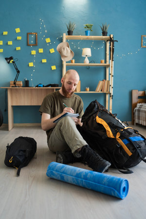 Thoughtful male traveler writing checklist of things for hiking trip at homeの写真素材