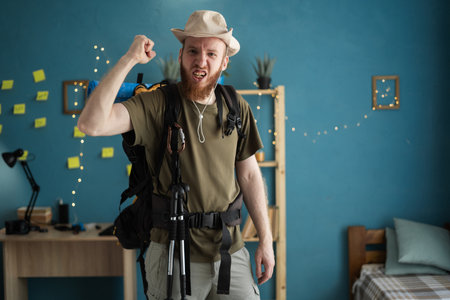 Young bearded man traveler in hat prepare for hiking vacation at home suffering angry with fist-up. Aggressive and angryの写真素材