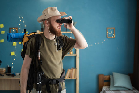 focused male traveler in hat with backpack looking through binoculars while standing at home,...