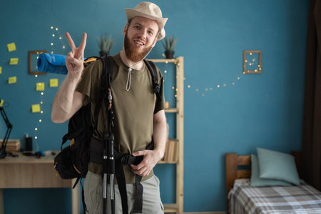 Young bearded man traveler preparing for hiking pointing two fingers victory gesture at home. Backpacking hikingの写真素材