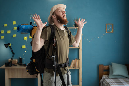 Young bearded man traveler preparing for hiking aggressive screaming at home interior. Backpacking hikingの写真素材