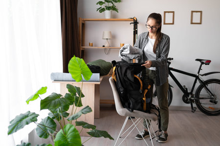 Female Backpacker in shirt and eyeglasses packing backpack with gear and clothes for her trip at...