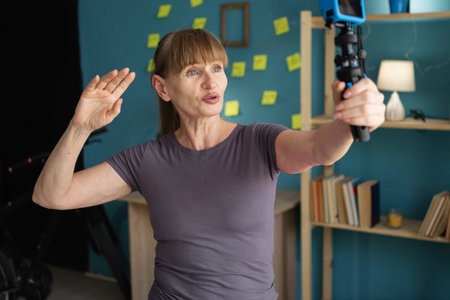 Middle aged woman in t-shirt making video call or taking selfie with modern action camera standing in living room at homeの写真素材