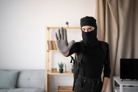 Female Burglar or thief wearing black balaclava stealing home showing stop gesture with palm of hand...
