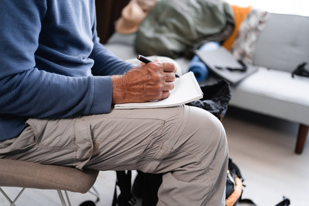 Close up of hands of elderly male traveler writing checklist list of necessary things for hiking and...