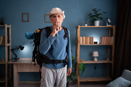 Retired traveler backpacker wearing hat with backpack preparing to trip looking at the camera and making a shh gesture at home. Trip, tourism, hiking and travelの写真素材