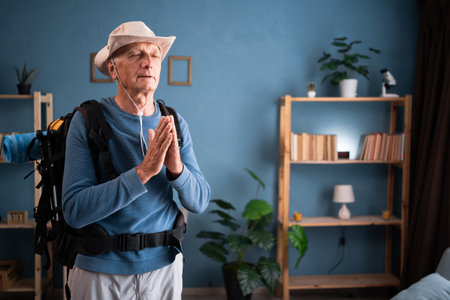 Meditation. Retired traveler with backpack stands in meditative pose, enjoys peaceful, hands in praying gesture at home interior. Tourist makes active healthy lifestyle. Hiking travel and tripの写真素材