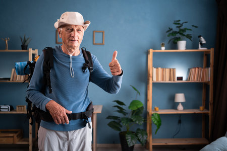Like gesture. Happy retired traveler backpacker man looking at camera show thumbs up like sign preparing to trip at home. Elderly grandfather hikerの写真素材