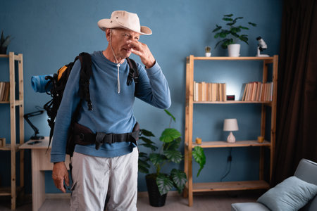 Elderly traveler with backpack pinching nose at home. Tourist likes active healthy lifestyle. Hiking, travel and tripの写真素材