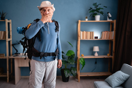 Shocked Elderly traveler with backpack with mouth covered at home. Tourist likes active healthy lifestyle. Hiking, travel and tripの写真素材