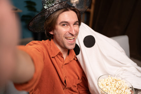 Happy witch man and scary ghost in halloween costume making selfie on smartphone sitting on sofa in the living room. concept of Halloween celebrationの写真素材