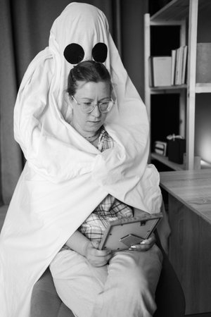 Concept of afterlife and memory for lost love. Sad woman looking old photo in frame sitting on chair while male ghost hugging her in the living room. Dead husband. Life and deathの写真素材