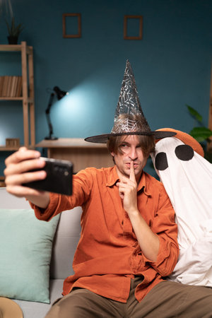 Happy witch man pointing shh gesture and scary ghost in halloween costume making selfie on smartphone sitting on sofa in the living room. Halloween celebrationの写真素材