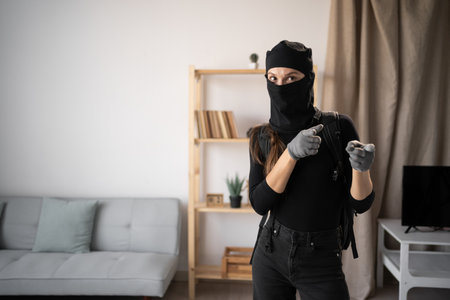 Burglar, robber and thief. Young woman wearing black balaclava and gloves pointing choosing you while into somebodys homeの写真素材