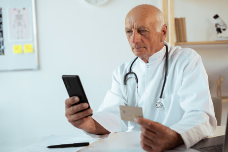 Elderly male doctor using smartphone and credit card for online payment. Online Banking in Clinicの写真素材