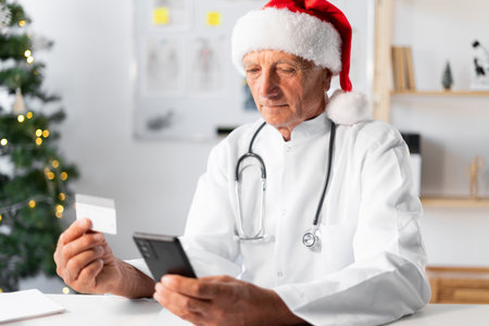Senior Elderly Doctor in Santa hat using credit card and mobile phone for online shopping on the shift in the hospital on Christmas Eve. medicine, holiday, technology, and healthcareの写真素材