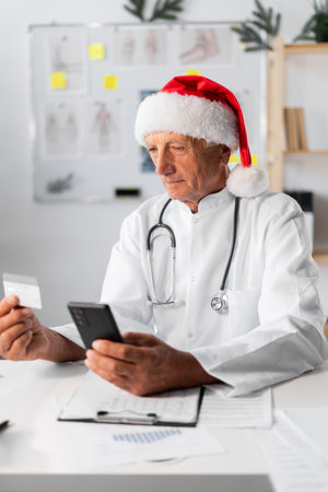 Senior Elderly Doctor in Santa hat using credit card and mobile phone for online shopping on the shift in the hospital on Christmas Eve. medicine, holiday, technology, and healthcareの写真素材