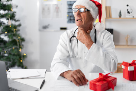 Senior elderly Doctor in Santa hat on the shift on christmas eve suffering sore throat from a cold. healthcare conceptの写真素材