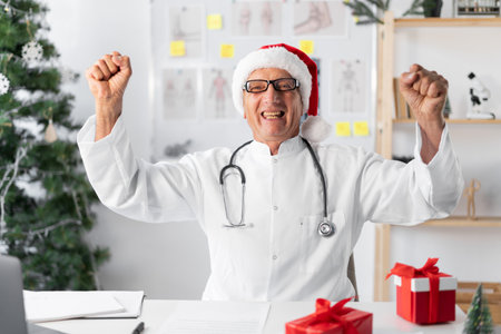 Senior elderly Doctor in Santa hat on the shift on christmas eve, winner and success fist celebrating good newsの写真素材