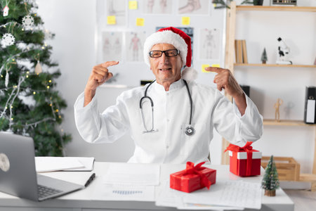 Senior elderly Doctor in Santa hat on the shift on christmas eve point index fingers on opened mouthの写真素材