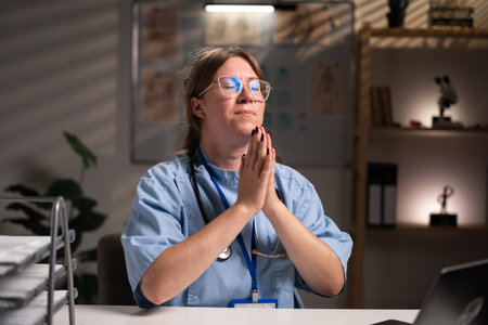 nurse working overtime on night shift holding hands folded, praying in hospital office. Health care and medicine serviceの写真素材