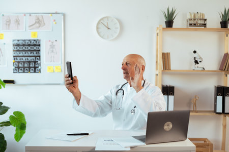 Elderly doctor holding phone making online video call. Male therapist patient consultation in telemedicineの写真素材