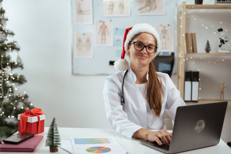 Portrait of smiling Female doctor in santa hat working online using laptop looking at camera on shift on christmas eve in the hospital. Holiday, medicine and healthcareの写真素材