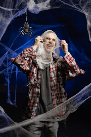 Person with scary Halloween zombie makeup on face standing covered in spiderwebs on blue background