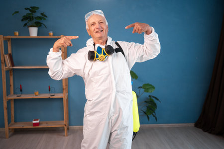 happy Elderly pest control worker point index fingers on opened mouth teeth inside infested house. Exterminator in protective suit holding spray bottle with insecticide for homeの写真素材