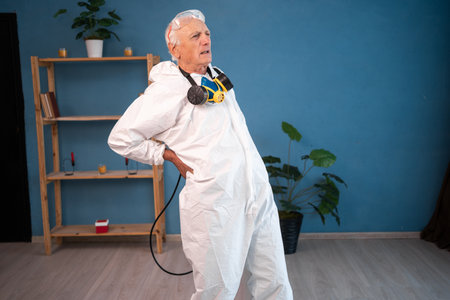 portrait of Elderly pest control worker suffering back ache and lower pain inside infested house. Exterminator in protective suit holding spray bottle with insecticideの写真素材