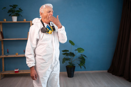 tired and yawning Elderly pest control worker inside house. Exterminator in protective suit holding spray bottle with insecticide for homeの写真素材