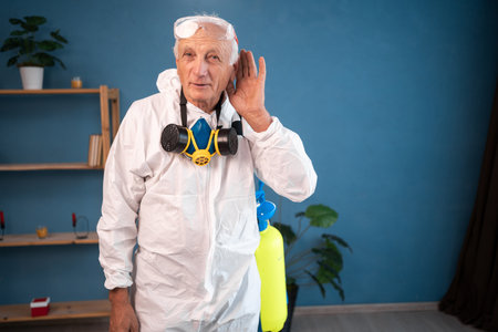 Elderly pest control worker with hearing problem listening inside infested house. Exterminator in protective suit holding spray bottle with insecticide for homeの写真素材