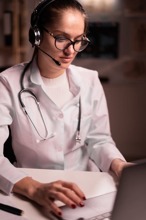 doctor in white coat and stethoscope during an online patient consultation working on night shift using laptop computer. Healthcare worker with headset giving virtual support. Telemedicineの写真素材