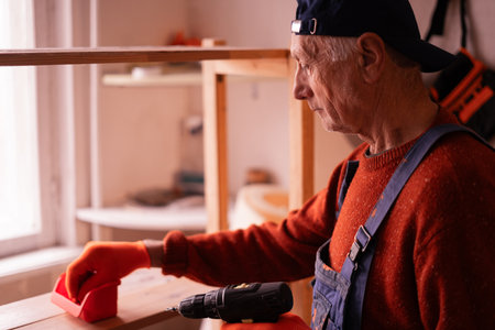 Senior Elderly Carpenter in blue overalls working in workshop. Craftsman assembling wooden shelf using electric drill in carpenter's shop, making furniture from wood. copy spaceの写真素材