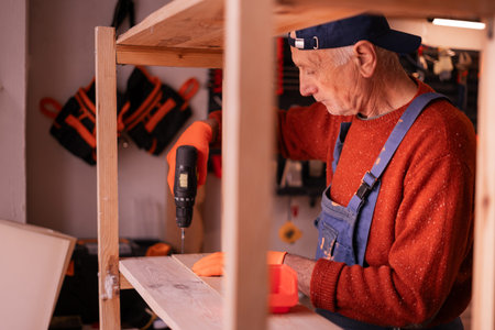 Senior Elderly Carpenter in blue overalls working in workshop. Craftsman assembling wooden shelf...