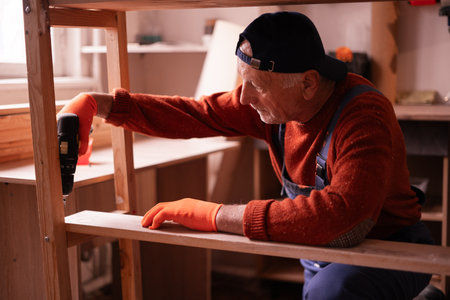 Profession, art and hobby concept. Elderly man carpenter assembling wooden shelf using electric bit drill working on wood in garage. Authentic workshop. copy spaceの写真素材