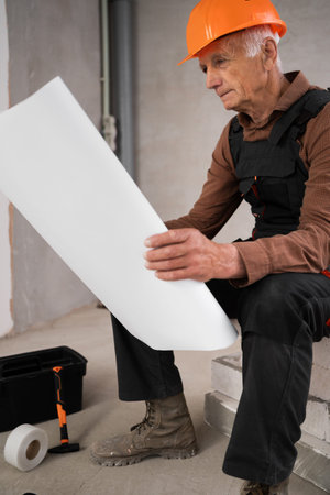 senior contractor in helmet and overalls reading paper blueprint showcasing expertise in...